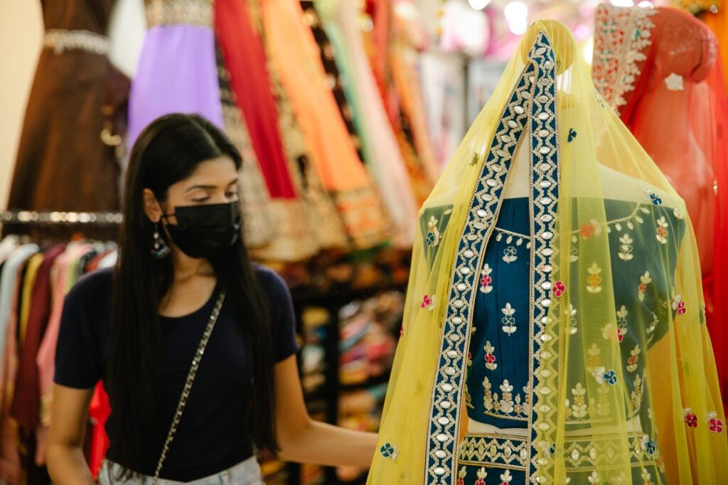 Woman wearing mask browsing traditional clothing in a vibrant marketplace.